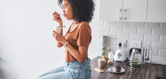 Shot of a young woman having a tub of yoghurt in the kitchen at home